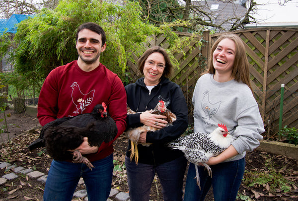 Abgebildet sind ein Mann und zwei Frauen, die Hühner auf dem Arm halten und in die Kamera lachen. Sie tragen ein Sweatshirt, eine Jacke mit Reißverschluss oder einen Oversize Sweatshirt mit dem Dein Gartenhuhn Motiv – ein Huhn aus einer feinen Linie.